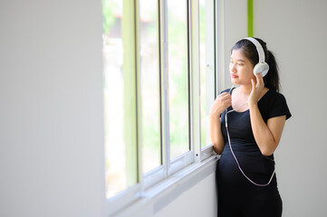 A pregnant woman in a black dress, wearing a white headphones, stood listening to music by the window.A pregnant woman standing by the window listening to nostalgic music