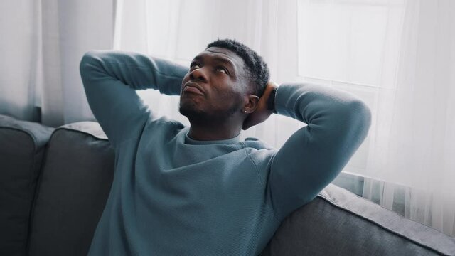 Serious man thinking while sitting on a blue sofa. Hands behind the head and looking at the ceiling of the room. Focused. Cozy room with white curtains in the background. 