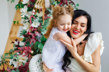 A woman with a little girl in beautiful white dresses in a bright studio decorated with flowers. Mother and daughter play, hug, have fun.