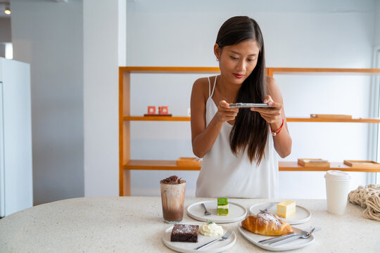 Asian woman using smartphone taking a photo of tasty cake dessert on white plate on the table. Female blogger influencer photographing sweet bakery and beverage for review in food restaurant blog