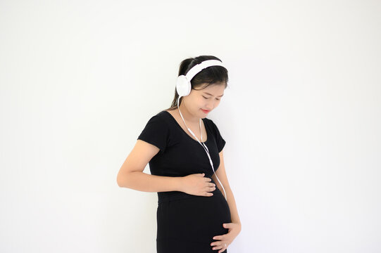 Pregnant woman wearing headphones wearing a black dress standing on a white background