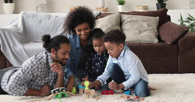 Full African young family with small son and daughter gather in living room play toy railroad enjoy weekend communicating spend quarantine time develop entertain kids use cool toys. Home hobby concept