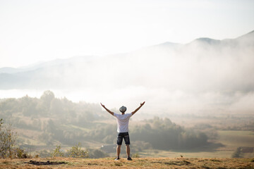 Fototapeta na wymiar Happy traveler man raising hands up on the top of the world above mountains in white clouds. Hipster guy enjoy amazing atmospheric moment. travel and wanderlust concept