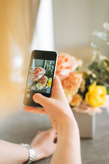 Stylish young woman shoots bouquet of flowers in box on mobile phone. Crop photo of hands using mobile