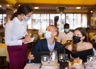 Positive man and woman in face masks for disease protection talking with waitress, choosing evening meal in restaurant, having date during coronavirus pandemic