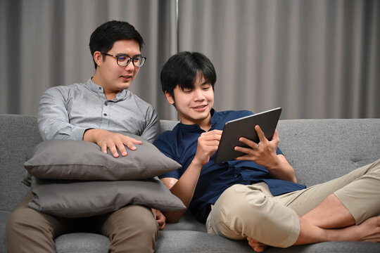 Casual homosexual couple using digital while sitting on sofa at home. Homosexual, LGBT concept. - Powered by Adobe