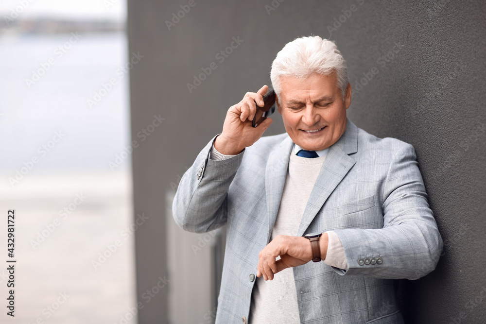 Poster Senior businessman talking by mobile phone near wall outdoors