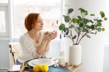 red haired woman in white sweater sits at table in cafe, holds mug of coffee or tea in her hands, and smiles, looks out window