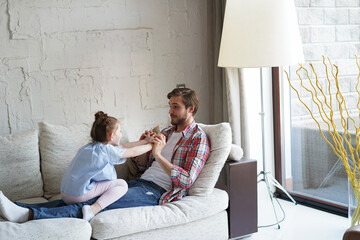 Cute little kid daughter laughing tickling playing with dad on sofa, happy father relaxing having...