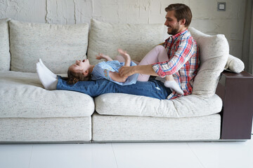 Cute little kid daughter laughing tickling playing with dad on sofa, happy father relaxing having fun with funny small child girl bonding enjoying leisure together in living room. - obrazy, fototapety, plakaty
