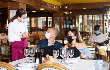 Fototapeta na wymiar Positive man and woman in face masks for disease protection talking with waitress, choosing evening meal in restaurant, having date during coronavirus pandemic