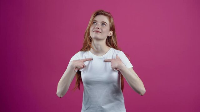 The young charismatic, confident redhead, points both fingers at her and says, me. Freckled teenage girl in a white T-shirt, isolated on a pink background. The concept of people's lifestyle