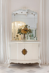 Old restored furniture in classic bright interior with white wall and curtains. Vintage mirror and flower bouquet on wooden cupboard