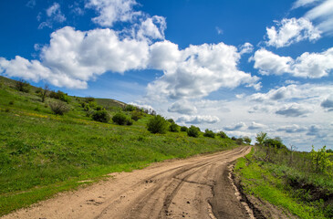 Dirt road at the foot of the mountain. A hillside with young greenery against the background of a sky with cumulus clouds. 
