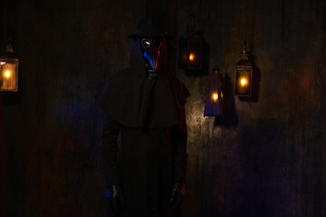 Fototapeta na wymiar Portrait of a plague doctor in a black leather mask, hat and hood, isolated against a dark wall with lanterns. Stylization of a historical costume, a creepy image, the concept of an epidemic.