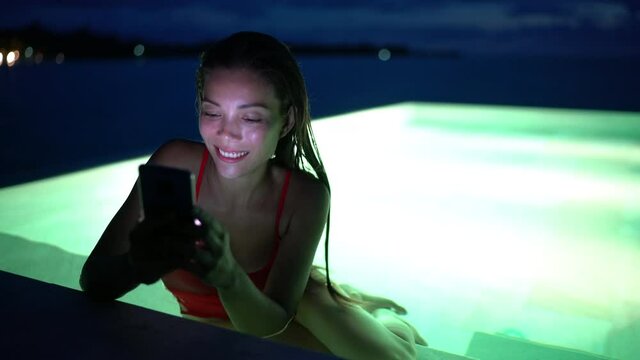 Smartphone - woman using mobile cell phone app on vacation in pool at night on travel holidays. Girl using smartphone app using phone app for banking, booking hotel and social media.