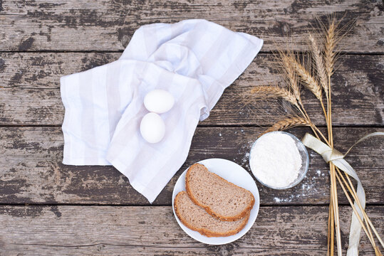 Two eggs on white napkin with spikelets of wheat, bowl of flour and bread on plate on wooden background table. Harvest season holiday World Bread Day. Bread is the staff of life. Top view.