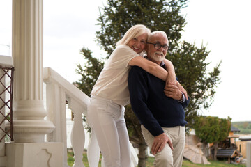 A good-looking senior couple is standing on a porch, talking and smiling. The wife stands on the higher step and holds her hands on a husband's shoulders.