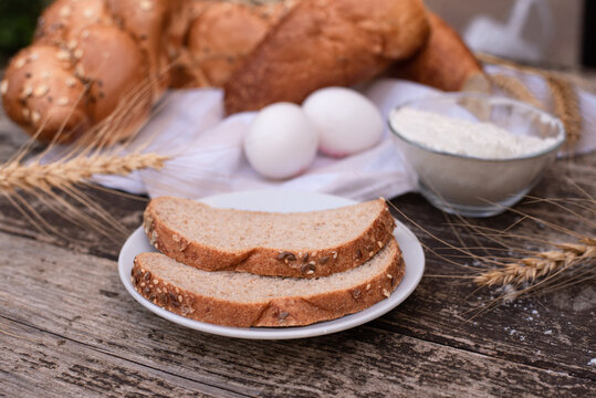 Fresh crispy bread on white napkin with spikelets of wheat, bowl of flour, for bread on plate and two eggs on wooden table. Harvest season holiday World Bread Day. Bread is staff of life. Side view.