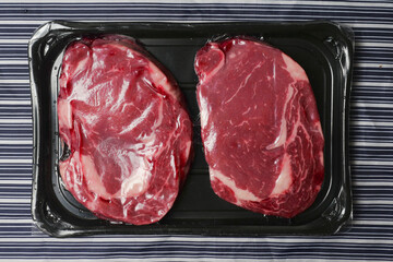 Fresh raw rib eye steak on a plastic tray on a black and white stripe butchers apron. Meat industry product. High quality beef with excellent marbling.