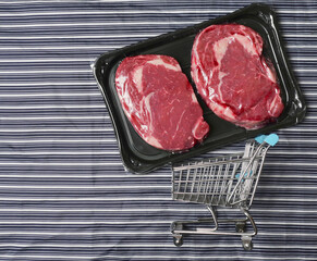 Fresh raw rib eye steak on a plastic tray and small shopping cart on a black and white stripe butchers apron. Meat industry product. High quality beef with excellent marbling.