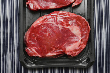 Fresh raw rib eye steak on a plastic tray on a black and white stripe butchers apron. Meat industry product. High quality beef with excellent marbling.