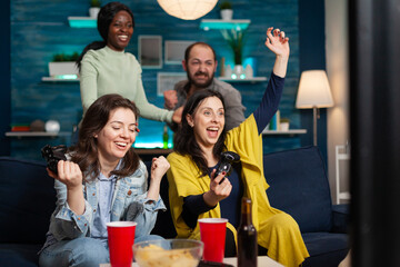 Cheerful women celebrating victory while playing video games with friends using wireless controller. Group of mixed race friends playing games while sitting on sofa in living room late at night.