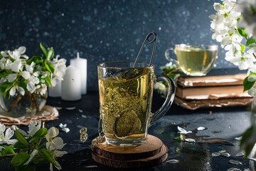 flower tea in a transparent mug on a dark background. Next to the cup are books and branches with white apple blossoms. The tea spills out of the mug