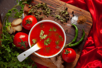 Close-up of Indian Homemade fresh and healthy tomato soup garnished with fresh coriander leaves and ingredients and herbs,  served in white ceramic bowl with spoon over red background. 