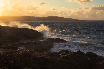 Waves crashing over rocks lit up with golden light from the setting sun on a beach in Qawra, Malta.