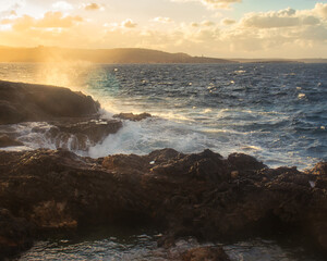 Obraz premium Golden light hitting water spraying over rocks on a beach in Qawra, Malta.