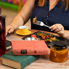 Woman working with computer and eating at dinner table. Business lady presses buttons on keyboard while holding fork with food in her other hand. Square format. Close up shot.