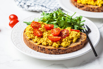 Tofu scramble toast with tomatoes and arugula on white plate, white marble background. Vegan food concept.