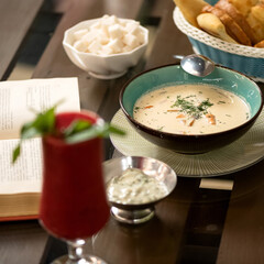 Reading while eating. Bowl of soup, glass of drink, and an open book on dining table. Square format. Close up shot. Soft focus.