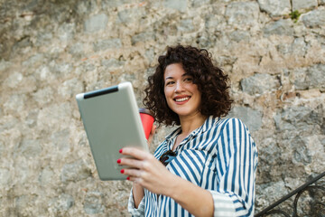 Attractive young woman with curly hair drinking coffee and reading her digital tablet while standing outside