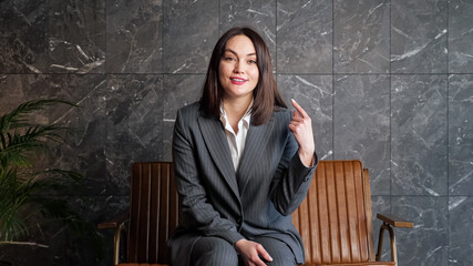 Smiling brunette in grey classic suit talks and gesticulates sitting on brown bench near large pot...