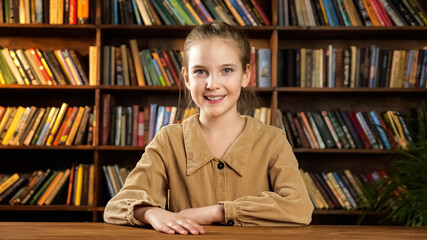 Nice schoolgirl model poses smiling cheerfully while sitting at new brown wooden desk against colourful books on racks and green pot plant