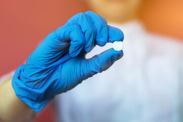 Fototapeta premium A young nurse, a doctor in blue rubber gloves, holds a white pill between her fingers. Close-up of an antibiotic in the hand for treatment. The concept of medical care, protection, rescue