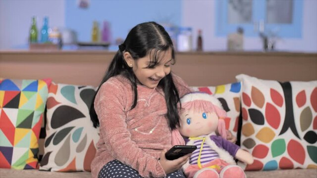 Lovely little girl using mobile sitting at her home - modern technology . Medium shot of a schoolgirl spending time with her doll and smartphone on a couch - new generation child