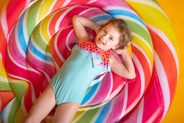 Happy little girl in swimming suit lying on colorful inflatable mattress looking up on yellow background.