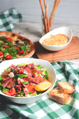 Prosciutto and arugula salad in a white bowl and cherry tomatoes pizza over wooden background. Red plaid tablecloth.