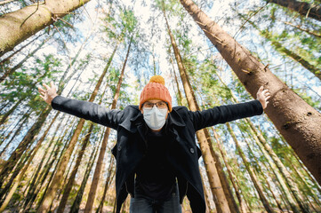portrait of attractive nerd in long sleeve black coat and orange hat with glasses and protective face mask outside in the pine forest  at sunset