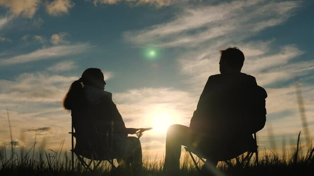 Happy couple on date admire beautiful sunset while sitting on travel chairs. Loving couple in park hold hands in sun. Family travels, rests on campsite. Happy people together nearby, enjoying nature