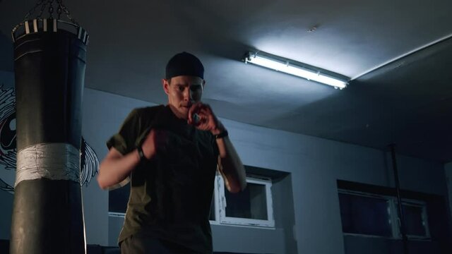 Alone boxer improves boxing movements in dark gym in slow motion. Young barefoot man training indoors on sport mats. Strong athlete in gym. Sport concept.