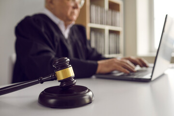 Close up of dark brown gavel on table on background of senior male judge working on laptop. Concept...