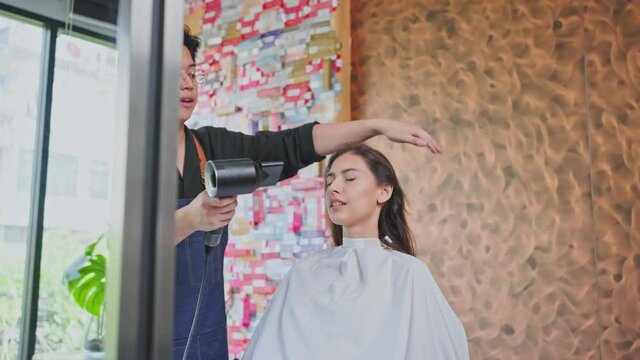 Asian hairdresser using hair dryer blowing customer's hair in salon.