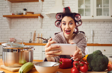 Crazy cheerful woman in bathrobe and hair curlers makes funny selfie with outstretched tongue on modern phone. Woman in a good mood having fun in the kitchen preparing a healthy dinner.