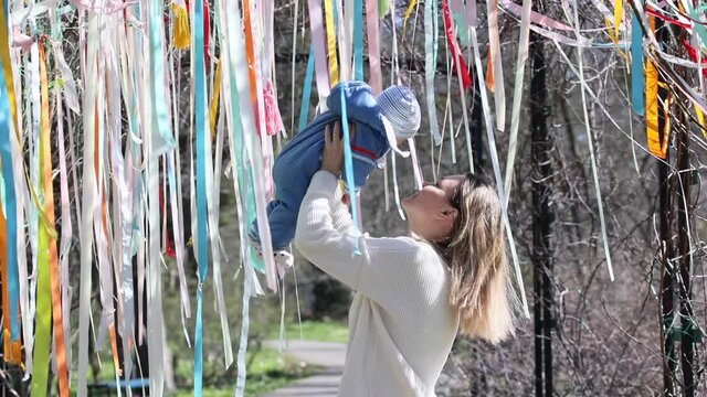 mother in white coat holding her infant baby in blue overall in the air and kisses him in the parc