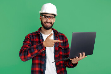 Smiling bearded engineer or constructor man in casual outfit pointing finger on computer over green background. - obrazy, fototapety, plakaty