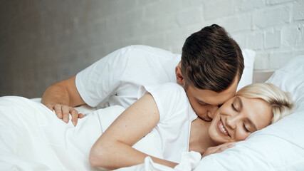 Fototapeta premium happy man kissing smiling woman lying with closed eyes in bed